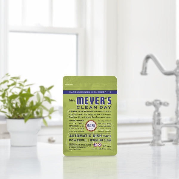 A green packet of Mrs. Meyers Automatic Dish Pacs sits on a white kitchen counter, offering a lemon verbena scent and sparkling clean with its oxygen-bleach formula. In the blurred background, a plant and faucet are visible.