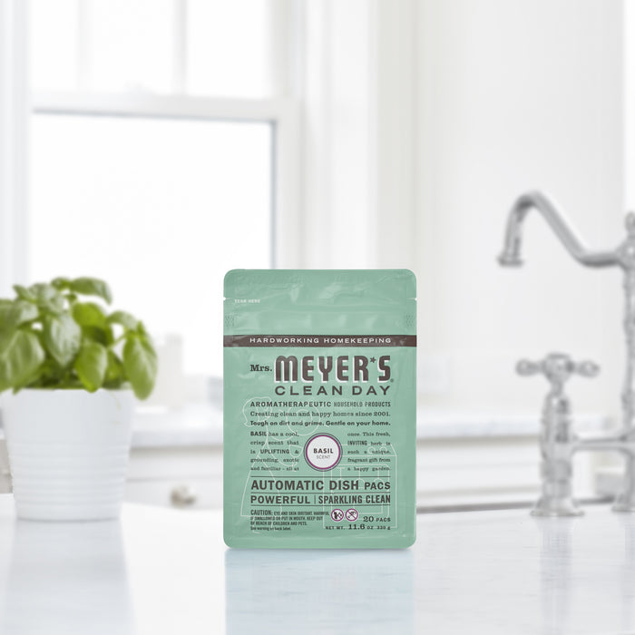 A pack of Mrs. Meyers Automatic Dish Pacs in basil scent rests on a kitchen counter near a potted plant, with light green packaging indicating its oxygen-bleach formula, a blurred kitchen faucet and sink in the background.