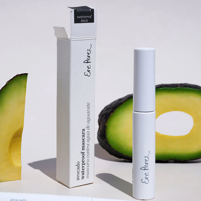 A white tube of Ere Perez Avocado Waterproof Mascara, a nourishing vegan option, stands by its waterproof black packaging. Two avocado slices are in the background against a light beige backdrop.
