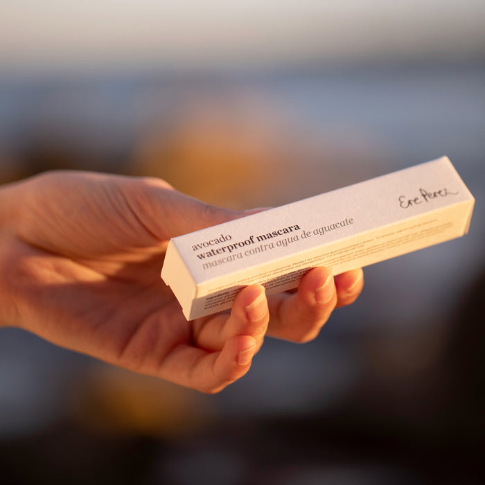 A hand holds a white box labeled Avocado Waterproof Mascara by Ere Perez against a sunset-hued outdoor backdrop. This nourishing, vegan mascara is beautifully packaged with bilingual text in English and Spanish.