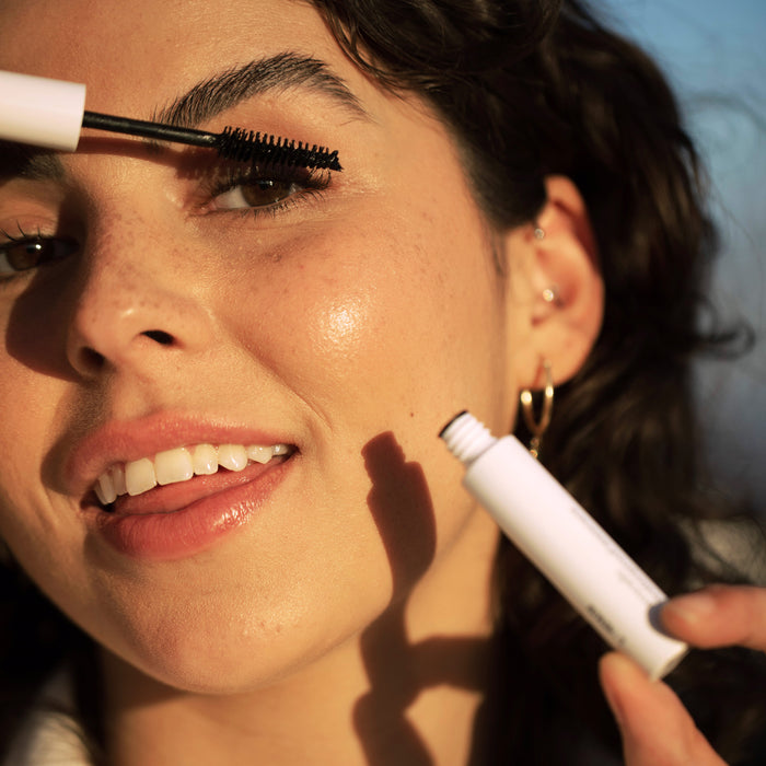 A curly-haired person applies Ere Perezs Avocado Waterproof Mascara with a brush to their upper lashes. They smile, adorned with multiple earrings, against an elegantly blurred background.