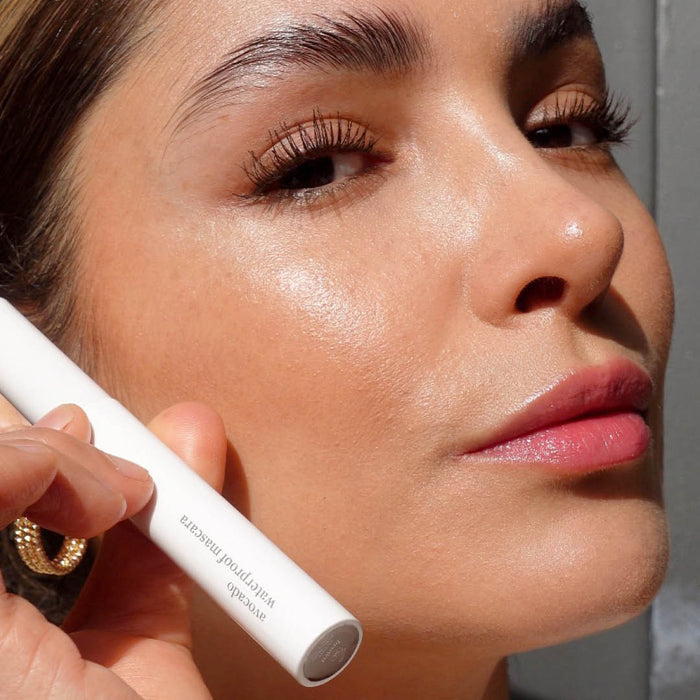 A woman with glowing skin holds a white tube of Ere Perez Avocado Waterproof Mascara near her face. Her brown hair is pulled back, and she has light makeup with glossy lips. Her earrings are partially visible, but the text on the tube isnt fully readable.
