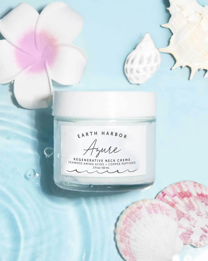 A jar of Earth Harbor AZURE Smooth Neck + Face Water Cream sits on a light blue surface with water ripples, encircled by a pink flower, white seashells, and pink and white clam shells. Infused with seaweed amino acids, it promises revitalization.