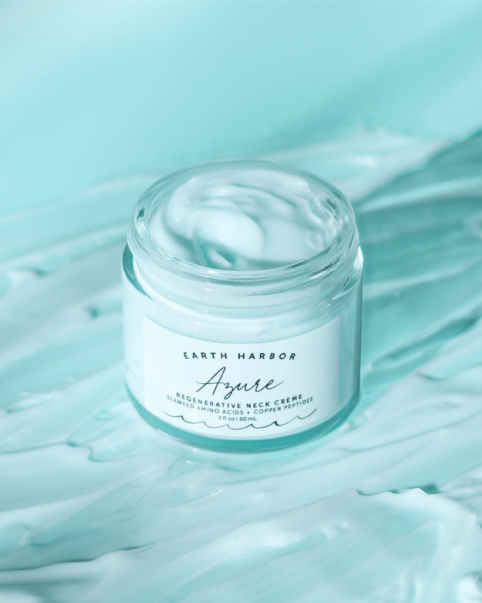 An open jar of Earth Harbor AZURE Smooth Neck + Face Water Cream rests on a teal surface. It reveals a light blue cream with Copper Peptides inside, set against a matching teal background that evokes a tranquil maritime vibe.