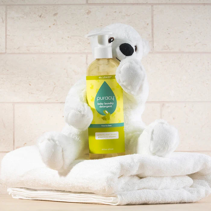 A white plush bear holds Baby Laundry Detergent while sitting on folded white towels.
