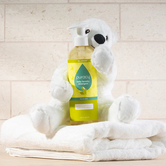 A white plush bear holds Baby Laundry Detergent while sitting on folded white towels.