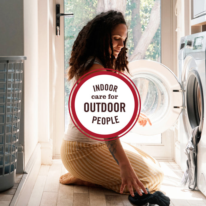 A woman kneels beside a washing machine, loading laundry with Mrs. Meyer’s Baby Laundry Detergent as sunlight pours through a glass door. The text overlay reads: “Indoor care for outdoor people.”.