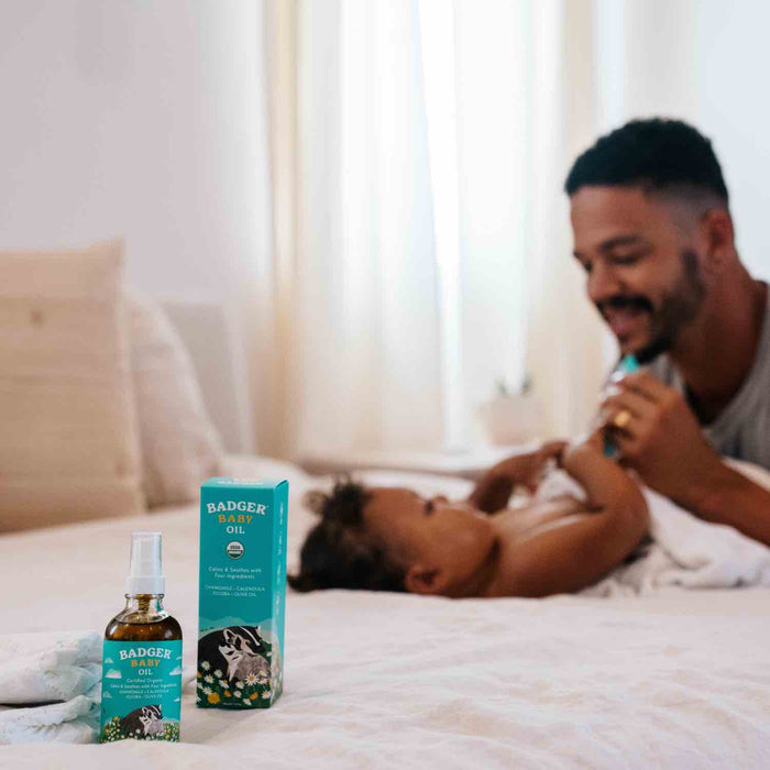 A baby lies on a bed, playfully interacting with an adult kneeling nearby. In the foreground are a bottle and packaging of Badger Balm Baby Oil, crafted with organic ingredients. Sunlight gently filters through a curtained window, creating a softly lit room.