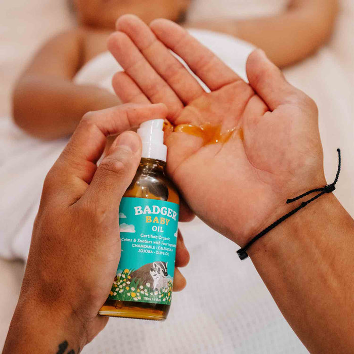 A person pours Badger Balms Baby Oil, enriched with organic ingredients, into their hand to apply it. In the blurred background, a baby lies on a white blanket. The oil label features a colorful badger design.