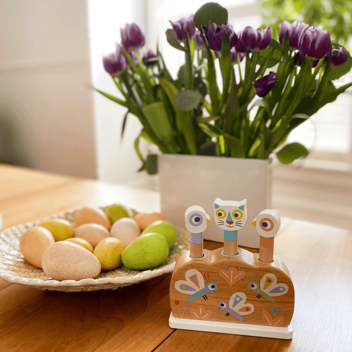 The DJECO Baby Popi Pop-Up Puzzle Toy, made from FSC® Certified Wood, sits decoratively beside a plate of pastel eggs. In the background, a vase holds vibrant purple tulips. Sunlight suggests how such toys enhance motor skills in an inviting setting.