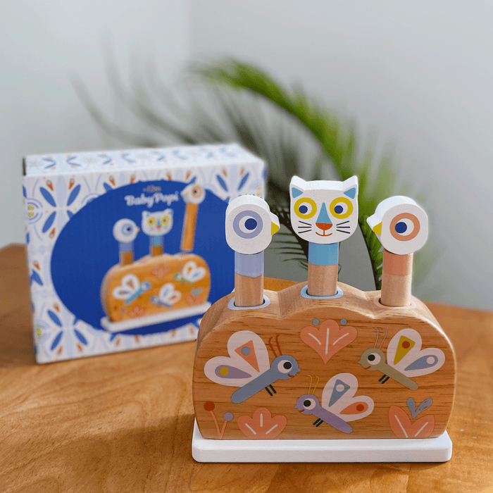 The Baby Popi Pop-Up Puzzle Toy by DJECO, made from FSC® Certified Wood, rests on the table. With animal heads and spring-loaded pegs, it aids motor skills development. The toy box displays colorful animal and insect illustrations while a leafy plant adds a natural touch in the background.