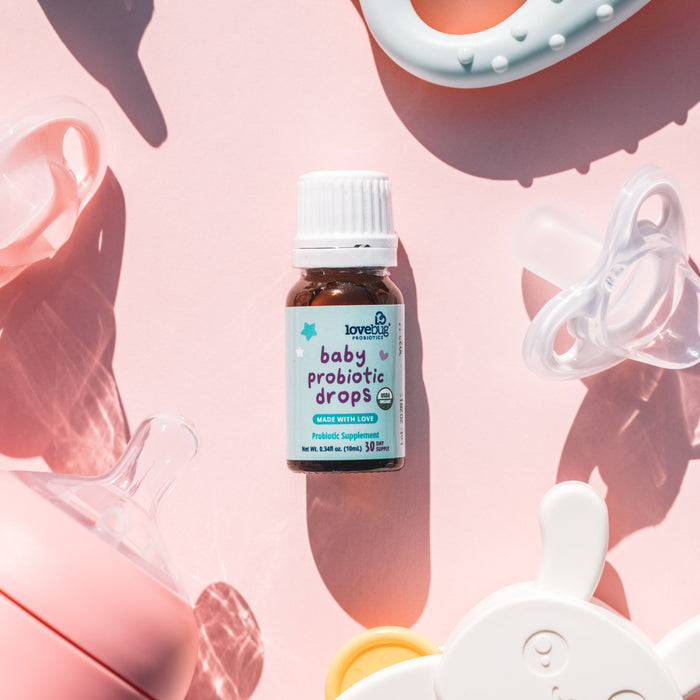 A small bottle of Lovebug Baby Probiotic Drops - Organic for digestive health sits on a pink surface, surrounded by pacifiers and a teething ring. Sunlight casts gentle shadows, highlighting the bottles role in bolstering your babys immune system.