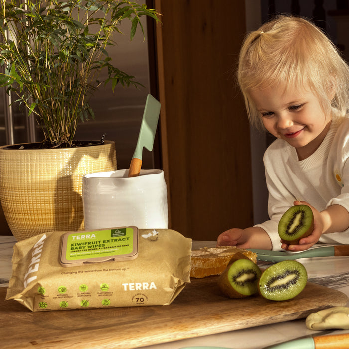 A young child smiles holding half a kiwifruit in the kitchen. On the counter are kiwis and Terra Baby Wipes - Kiwi Fruit, known for their natural moisturizing properties. A potted plant and a white utensil container complete the cheerful scene.