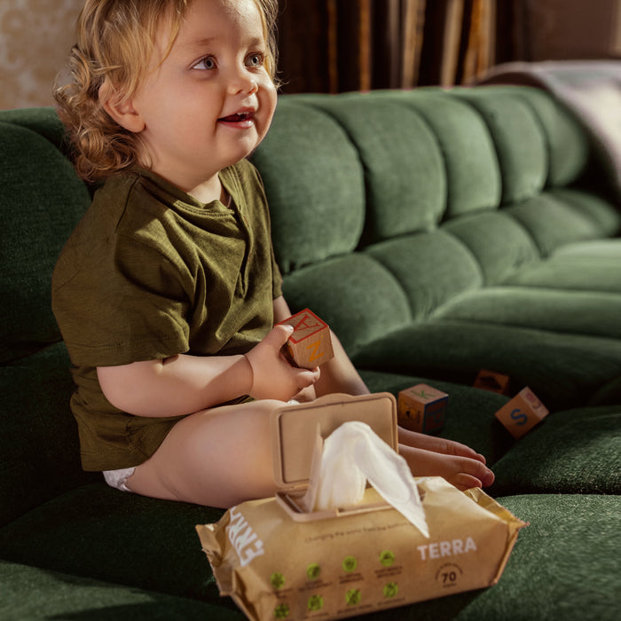 A child in a green shirt sits on a green couch, holding a wooden block, with an open pack of Terras Baby Wipes - Kiwi Fruit beside them. More wooden blocks are scattered around.