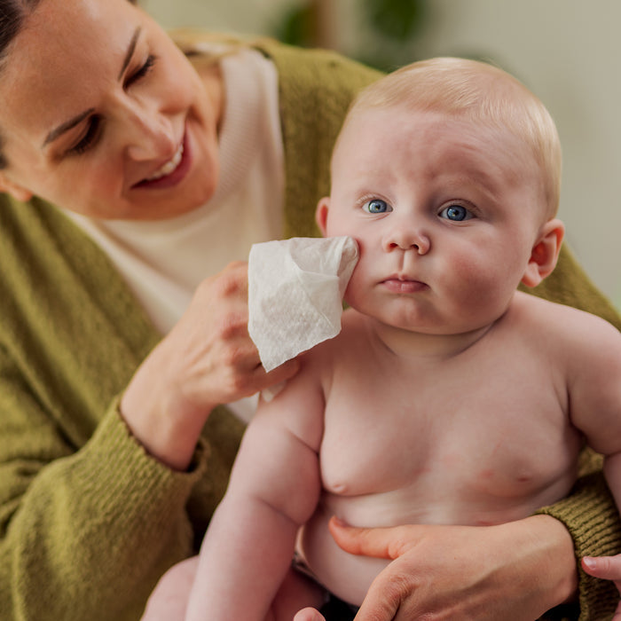 A smiling woman in a green sweater gently wipes the cheek of a serious, shirtless baby on her lap with DYPER Baby Wipes for Sensitive Skin.