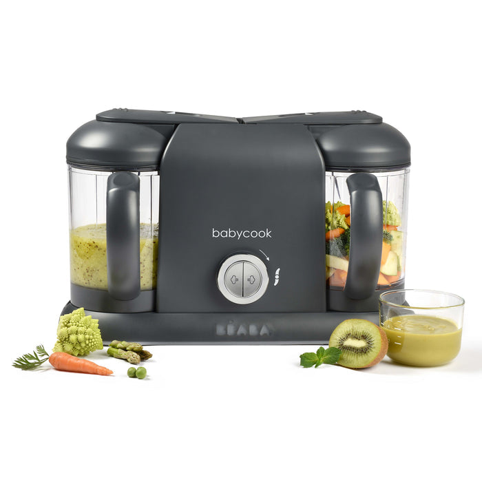 The BEABA Babycook Duo Baby Food Maker in gray features one-handed operation and two clear compartments filled with vegetables. In front, assorted veggies and a glass of homemade food highlight the convenience of preparing fresh meals.