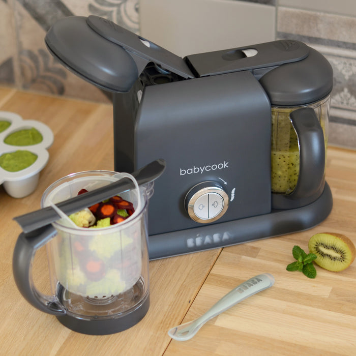 A gray BEABA Babycook Duo sits on a wooden countertop, ideal for making homemade baby food with one-handed use. One container has green puree, the other vegetables and fruit. Nearby are a tray of green cubes, a kiwi slice, mint leaves, and a spoon.