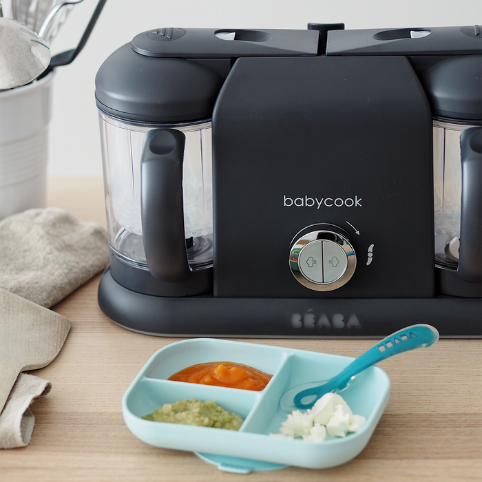 On a wooden countertop sits a sleek black BEABA Babycook Duo Baby Food Maker, ideal for homemade baby food with one-handed use. In front, a teal plate cradles portions of orange and green purees alongside white mashed food, complemented by a matching spoon.