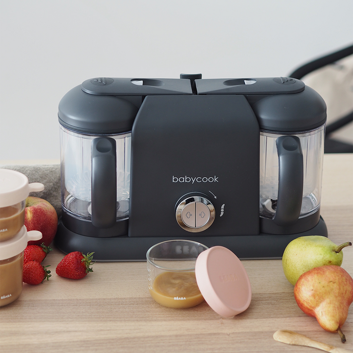 The BEABA Babycook Duo sits on the kitchen counter, ideal for homemade baby food. With dual compartments and one-handed operation, its surrounded by fresh strawberries, a pear, a banana, and jars of pureed delights with lids.