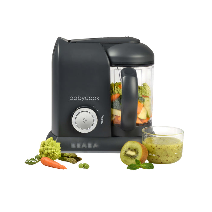 The BEABA Babycook Solo Baby Food Maker in black, featuring steam cooking for nutritious meals, has a transparent container with chopped vegetables. In front, there’s a glass of green homemade baby food next to kiwi slices, broccoli, carrots, and peas.