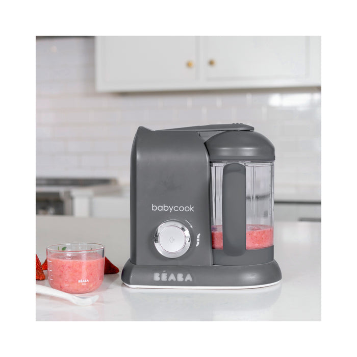 A gray BEABA Babycook Solo Baby Food Maker with a transparent container sits on the counter, ideal for steam cooking. A glass bowl holds a pink mixture and cut strawberries on a white plate in front of a blurred kitchen background, perfect for homemade baby food enthusiasts.