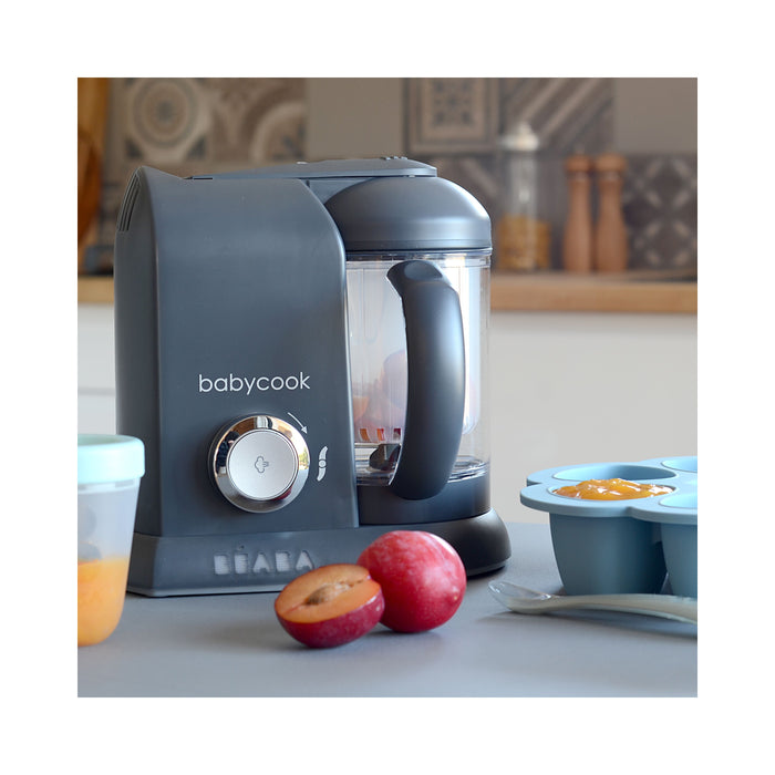 A gray BEABA Babycook Solo Baby Food Maker sits on the counter, ready to steam cook homemade baby food. Nearby, a halved plum rests beside containers of puréed food and a spoon, all set against a softly blurred kitchen background.