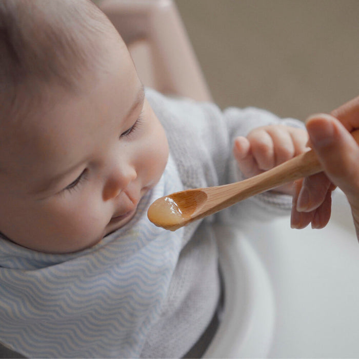 A baby in a high chair, wearing a bib, is being fed with Bambus Babys Feeding Spoon (6M+). The person feeding them holds the spoon near the babys mouth, offering a small amount of pureed food.