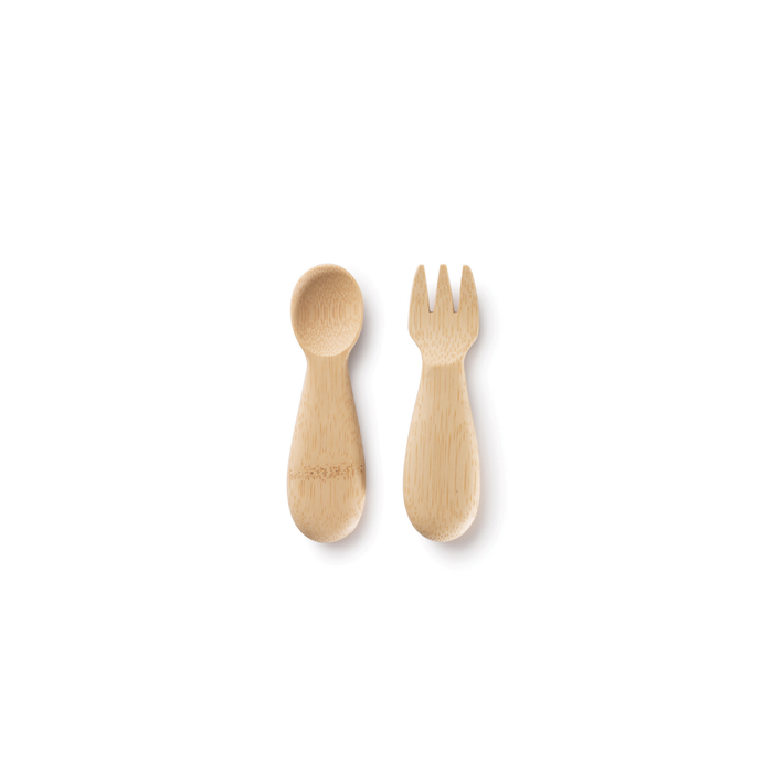 On a white background, two bamboo utensils from Bambus Babys Fork & Spoon set (12M+); the left is an organic spoon with a rounded handle, while the right is a three-pronged fork. Both have a natural wood finish, making them perfect for any toddler feeding set.