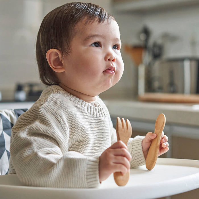 A toddler in a beige sweater sits in a high chair, clutching Bambus Babys Fork & Spoon (12M+). The child gazes slightly upward against a blurred kitchen background with countertops and appliances.