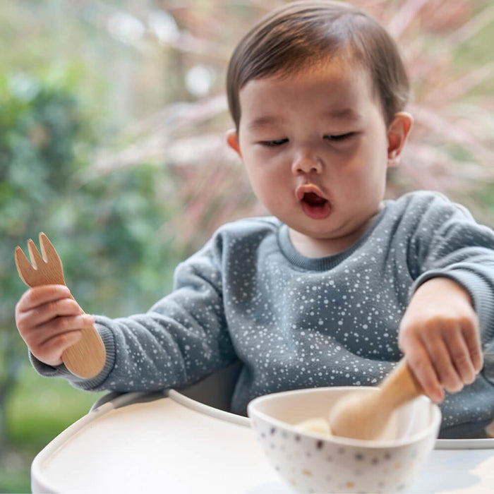 A toddler sits in a high chair near a window, clutching a Bambu Babys Fork & Spoon (12M+). Dressed in a gray, speckled sweater, the child concentrates on the bowl in front of them, while blurred greenery is visible outside.