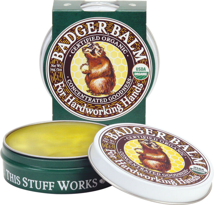 An open tin of Badger Balm for Hardworking Hands displays its yellow balm, featuring packaging with a badger illustration and phrases like Certified Organic and This Stuff Works! Made with organic ingredients, a closed tin is seen in the background.