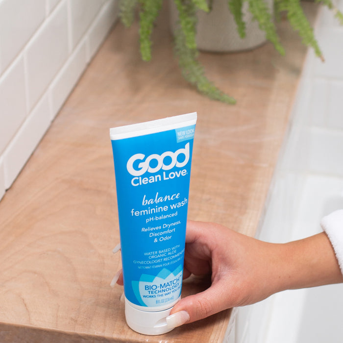 A hand holds a tube of Good Clean Love Balance Moisturizing Wash on a bathroom counter with tiles and a plant. The packaging emphasizes its Bio Match™ formula for relief from dryness, discomfort, and odor with pH balance.