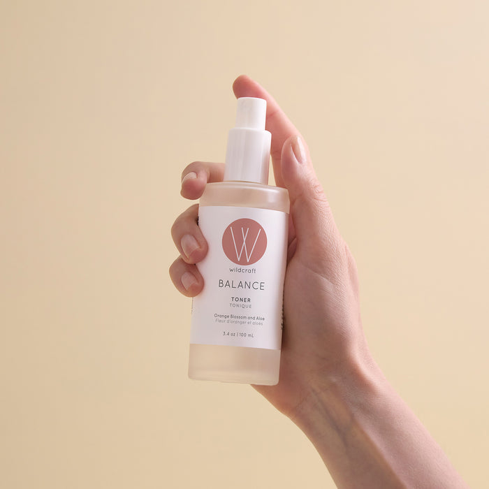 A hand holds a transparent spray bottle of Wildcraft Balance Toner, featuring a white label with a pink logo, set against a plain beige background.