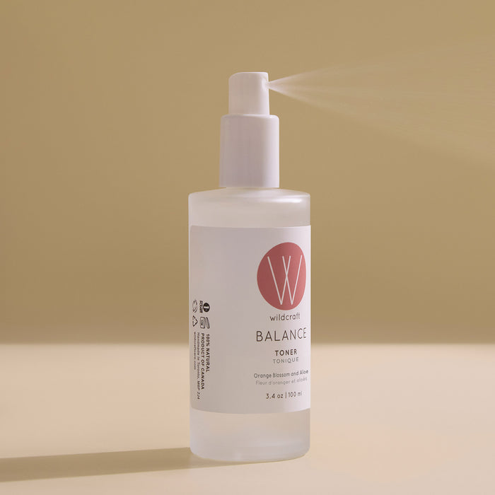 A clear frosted bottle of Wildcraft Balance Toner with a white pump sprays a fine mist. The hydrating toner with neroli and aloe vera sits on a soft beige background, its white label featuring a pink logo and black text.