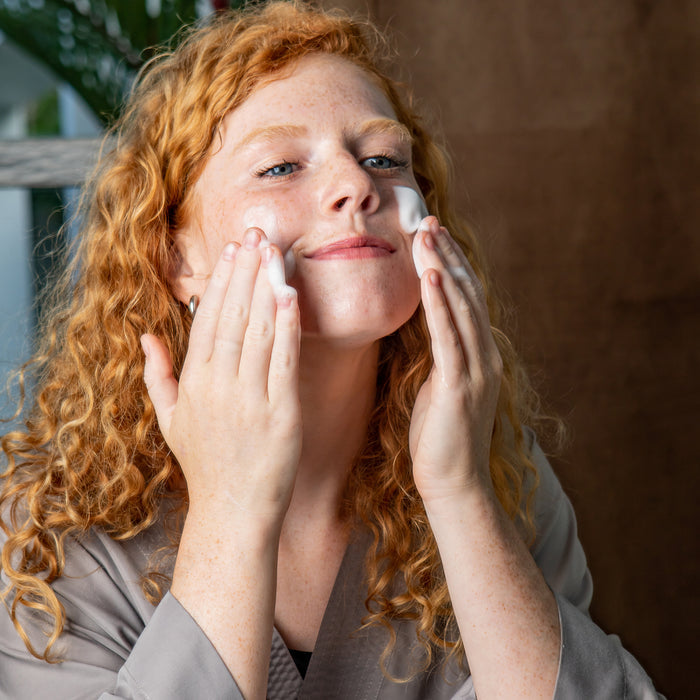 With curly red hair, a person uses both hands to apply Tree To Tubs Balancing Tea Tree Face Wash for Oily Skin, smiling slightly in a gray robe. The scene is set with a plant and brown fabric curtain, perfect for their skincare routine.
