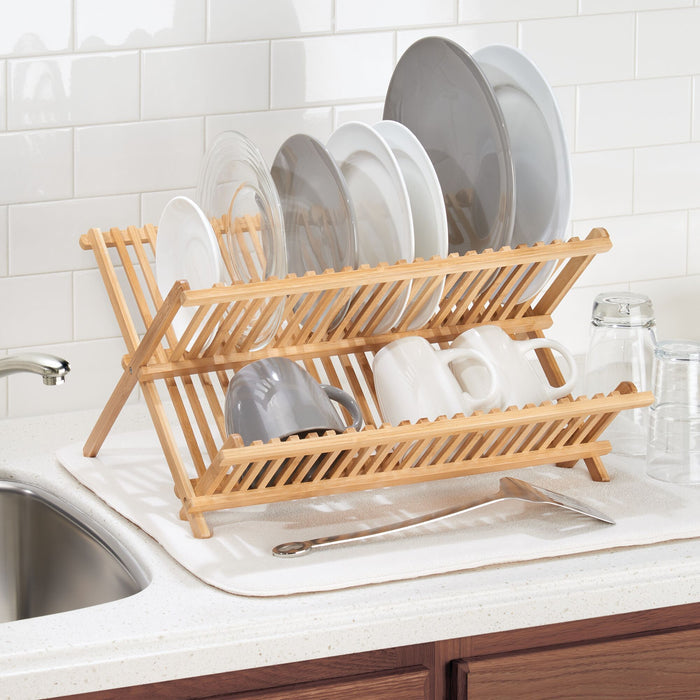 On the kitchen counter rests an iDesign Bamboo Folding Collapsible Dish Drying Rack, gracefully placed next to a shiny chrome faucet over a white sink. Its design effortlessly holds plates and cups, complemented by light-colored counters and a white tile backsplash.
