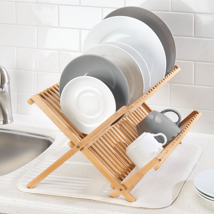 The iDesign Bamboo Folding Collapsible Dish Drying Rack neatly stacks white and gray plates and cups on a countertop by the sink. Set against white subway tiles, its renewable bamboo construction and collapsible design blend elegance with practicality.