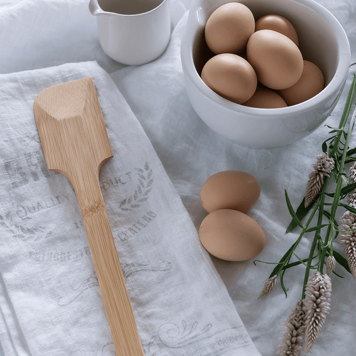 A Bambu Scraping Spatula rests on a white cloth next to a bowl of brown eggs, with two eggs beside it. A floral sprig lies nearby, and a ceramic cup in the background completes the eco-friendly kitchen vibe.