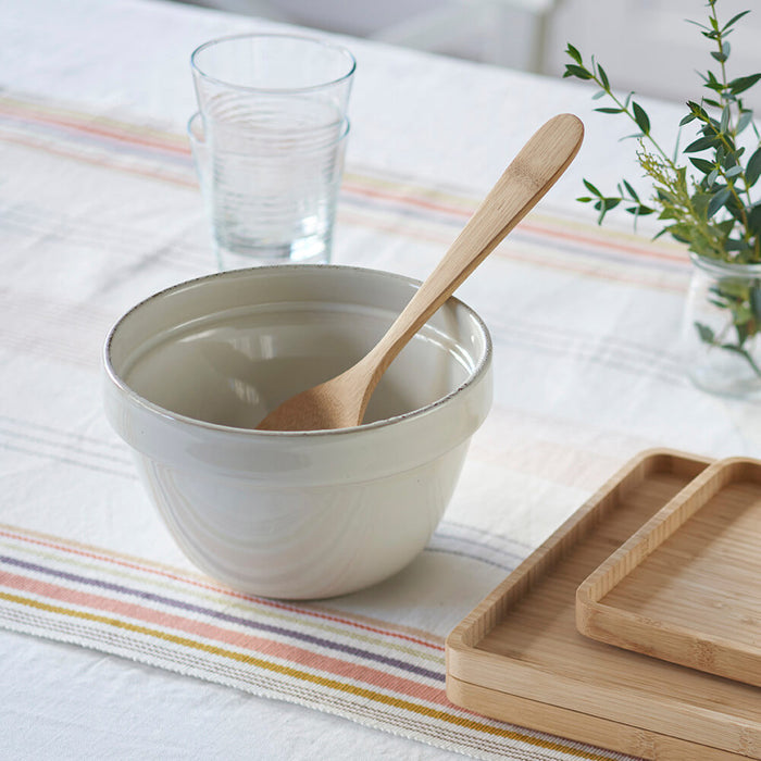 A ceramic mixing bowl with a Bambu serving spoon rests on a striped tablecloth. A drinking glass and wooden tray are close by, while a small plant in the background adds greenery.