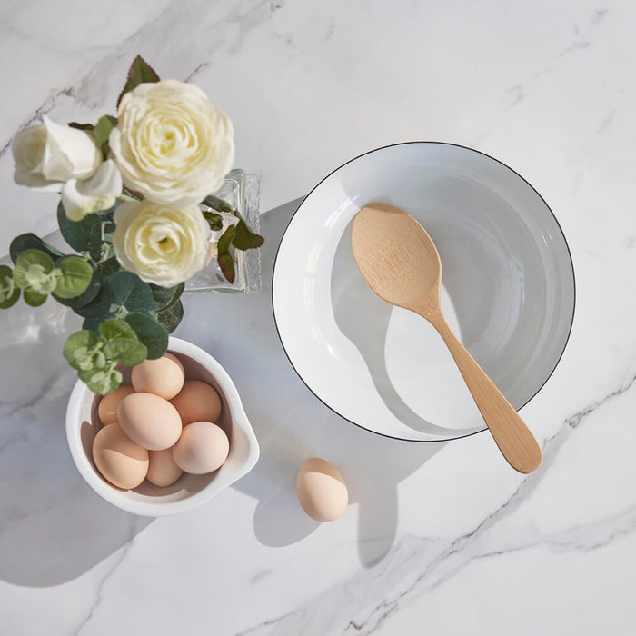 A white bowl with a Bambu Serving Spoon sits on a marble countertop. Nearby, a smaller bowl holds brown eggs, with one placed outside. A vase of white roses and greenery completes the serene setting.