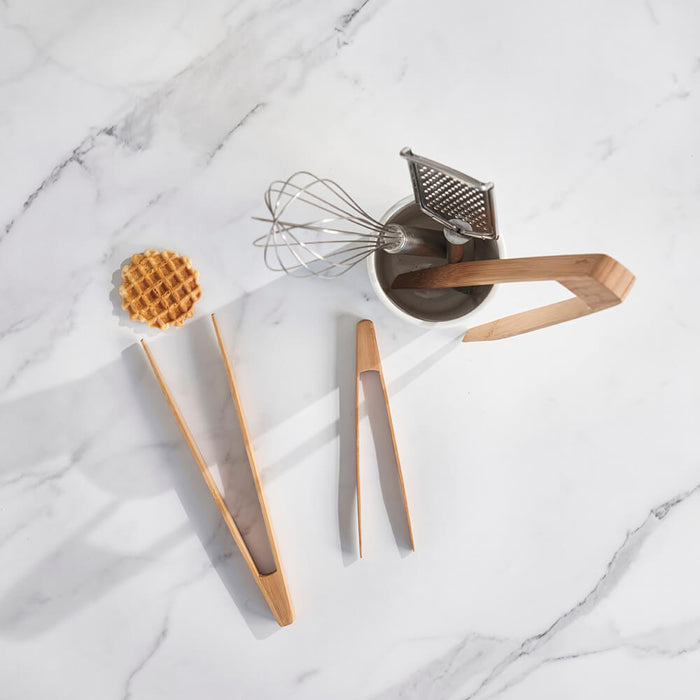 On a marble surface, Bambu Large Tongs hold a waffle next to smaller wooden tongs, an elegant metal whisk, and a grater in a cup—ideal for preparing outdoor grilling delights.