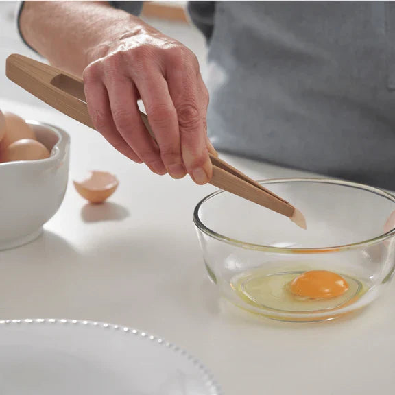 A person uses Bambu Bamboo Tongs, Large to pick up an eggshell over a glass bowl with a cracked raw egg, next to whole eggs and eggshells on a white countertop.