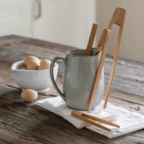 A ceramic pitcher holds Bambu's Bamboo Tongs, Large, on a white cloth atop a rustic wooden table, with a bowl of eggs and some loose eggs nearby.