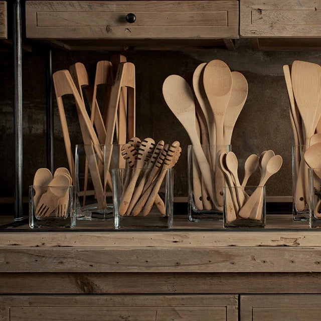 The Bambu Bamboo Tongs, Large, are displayed upright in a clear glass container on a rustic wooden shelf with drawers, keeping your kitchen organized—perfect for cooking or outdoor grilling.