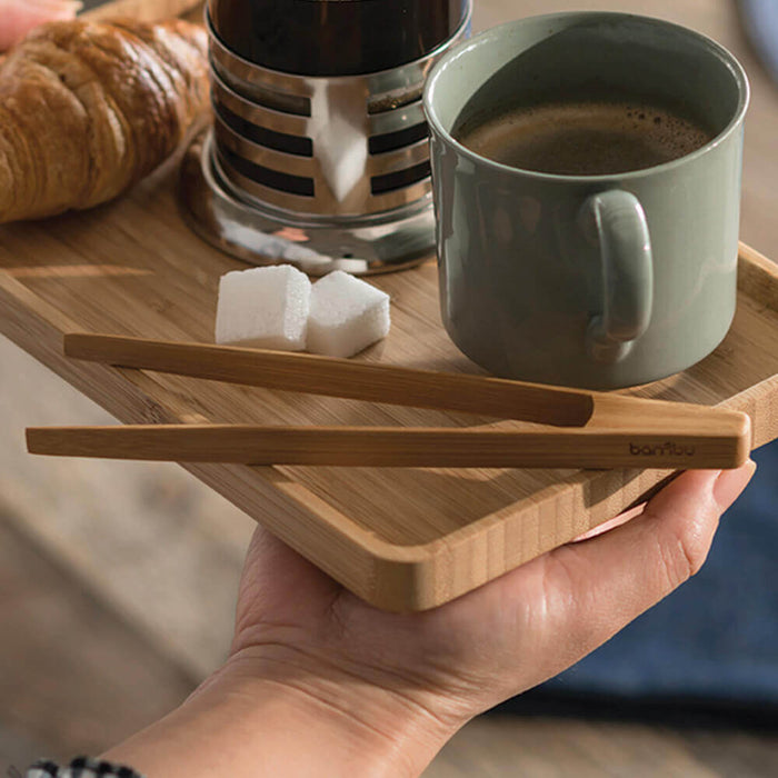 A hand holds a wooden tray with a green mug of coffee, a metal container, two sugar cubes, Bambus Small Tongs made from organic bamboo, and a croissant. Natural light brightens the cozy setting.
