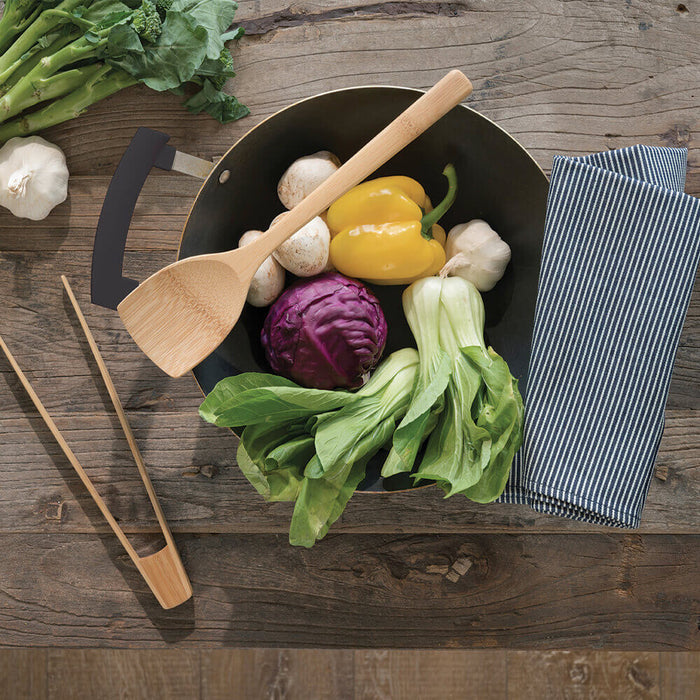 A wooden table features a large black pan filled with bok choy, yellow bell pepper, red cabbage, and mushrooms, ideal for Asian cuisine. A Bambu Wok Spatula is nearby, alongside garlic and a striped napkin. Wooden utensils are positioned on the left.