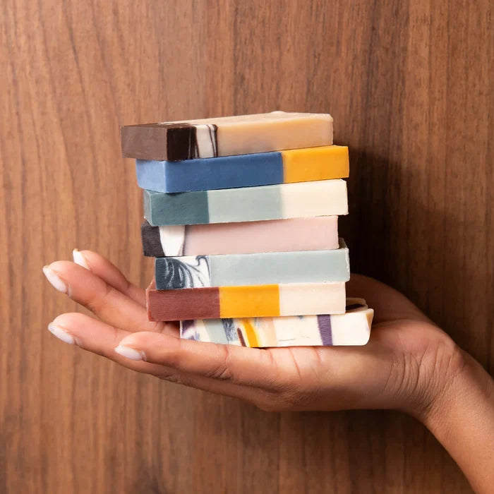 A hand holds a stack of seven colorful Pacha Peaces bar soaps by Pacha Soap Co. against a wooden background, displaying the vibrant variety pack.