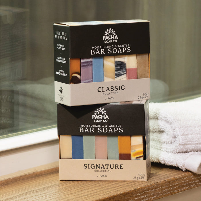 Two boxes of Pacha Soap Co. Bar Soap, Pacha Peaces, labeled Classic Collection and Signature Collection, rest on a wooden surface by a towel and window. Each variety pack features seven moisturizing, colorful bars.