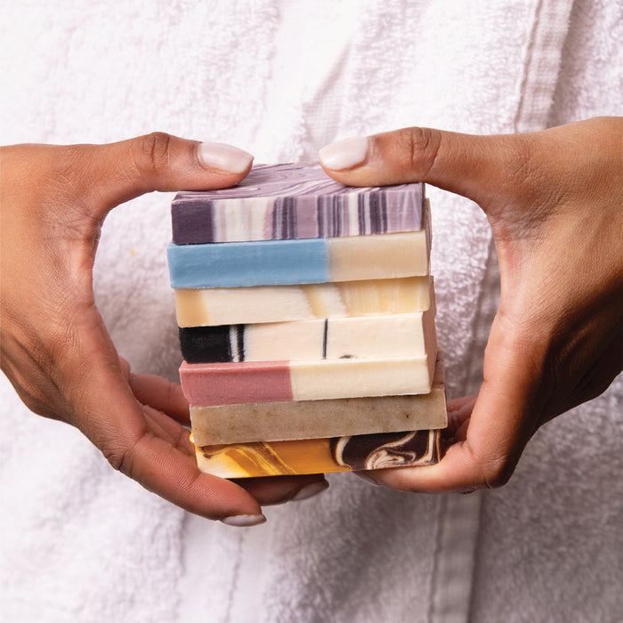 A person in a white robe holds six colorful, patterned Pacha Peaces Bar Soap bars by Pacha Soap Co. with both hands against a neutral background.
