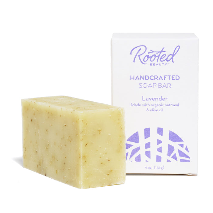 A beige handcrafted moisturizing soap bar with visible oatmeal bits sits beside its white box labeled Rooted Beauty Bar Soap, Lavender. Featuring purple text and designs, its crafted cruelty-free with organic oatmeal and olive oil.